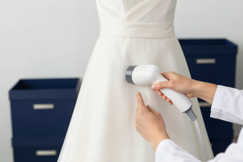 Illustration: a garment-care specialist’s hands steaming the skirt of an ivory wedding gown in a bright workroom
